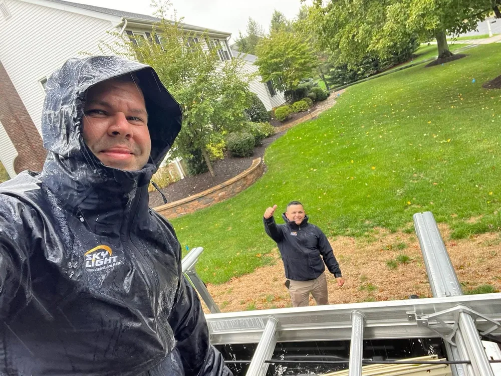 A Skylight Roofing inspector in wet weather gear at the top of a ladder during rain