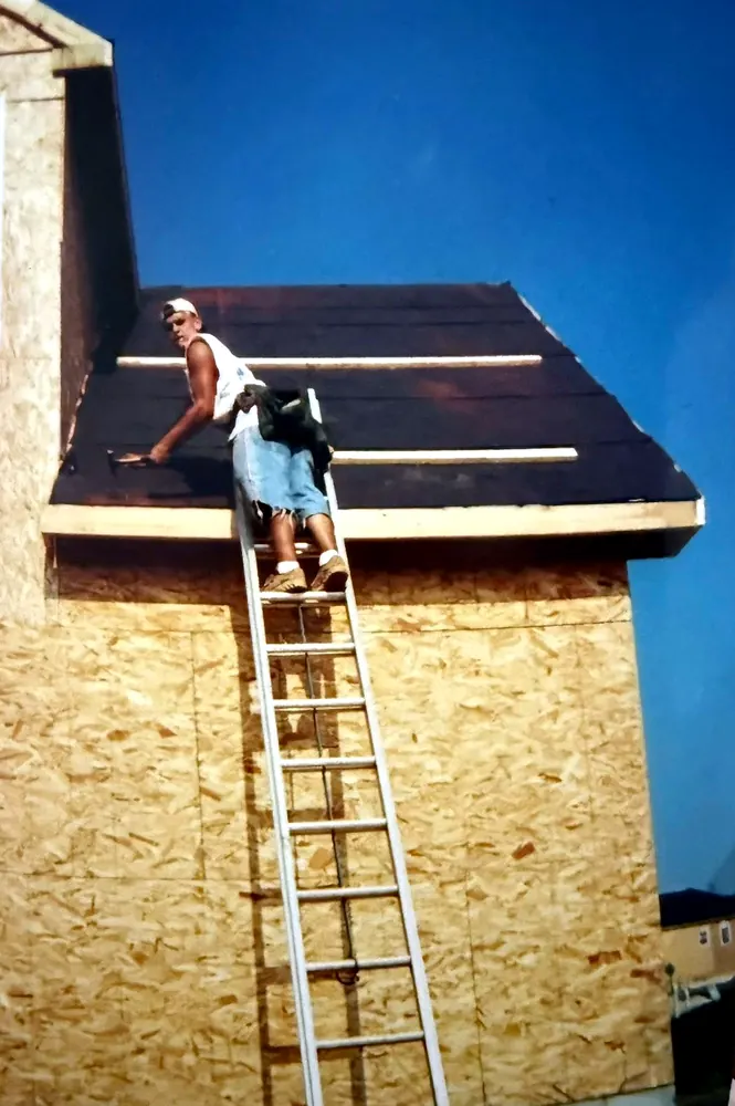 Mauricio Saenz as a young roofer early in his career, kneeling on a stripped roof deck laying underlayment, with an extension ladder up to the eaves