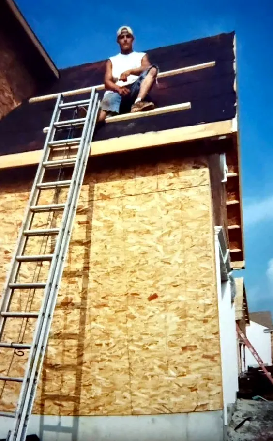 A young Mauricio Saenz sitting on the edge of a half finished roof, above a house still in bare sheathing